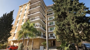 Um edifício residencial com várias andares, varandas, cercado por árvores e carros estacionados.