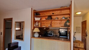 A compact kitchen with wooden cabinetry, a built-in oven, and a microwave. There is a wooden chest of drawers and a wicker chair.