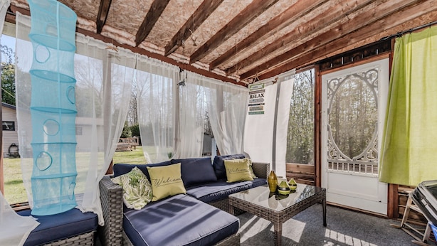 A covered outdoor seating area with wicker furniture, a glass table, and a view of the forest. - By The Beach - Affordable Sauble Beach Getaway (Sauble Beach)