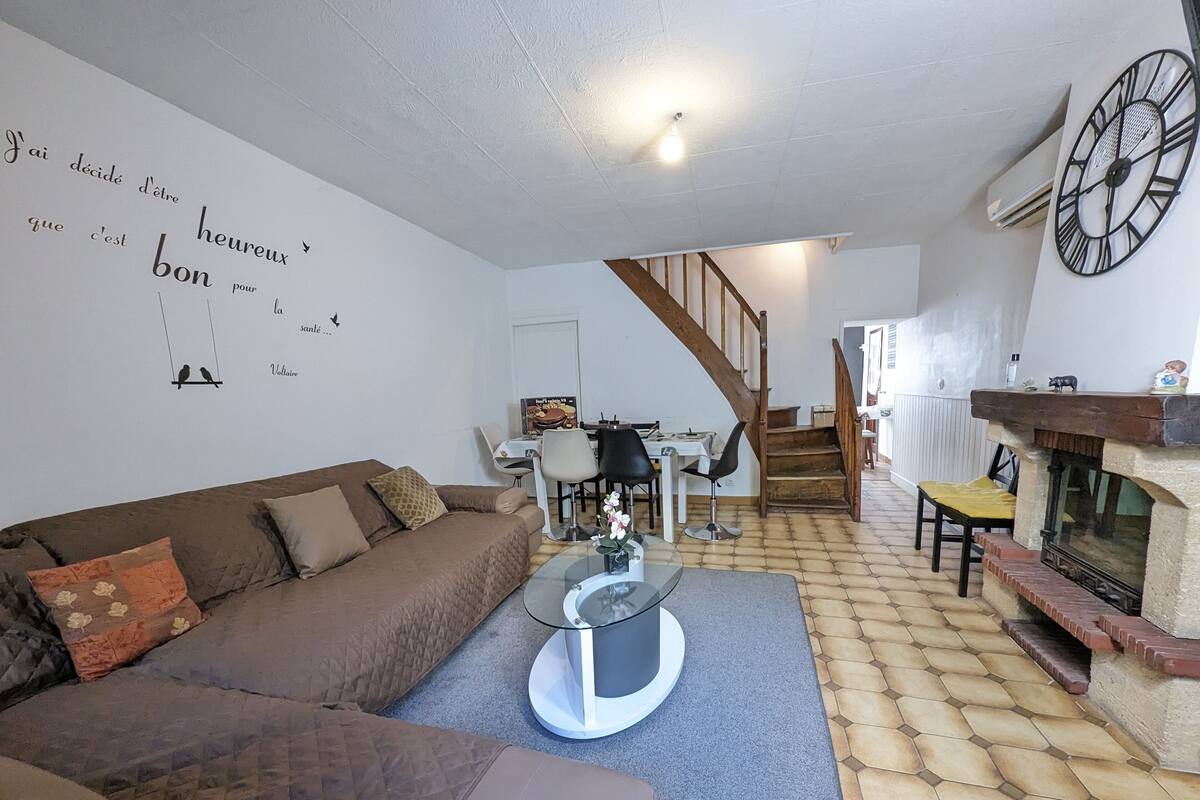 Een gezellige woonkamer met een bruine bank, een glazen salontafel en een open haard. Een trap leidt naar een andere kamer.