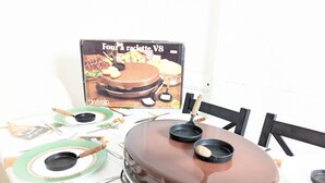 A dining table with a chocolate cake, a box labeled "Four à raclette V8", and a menu titled "Mon joli jardin".