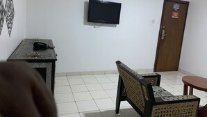 A room with a television mounted on the wall, a wooden door with a sign, a patterned table, a woven chair, and a small round table. - FOSTER INN -ASAKE (Ibadan)