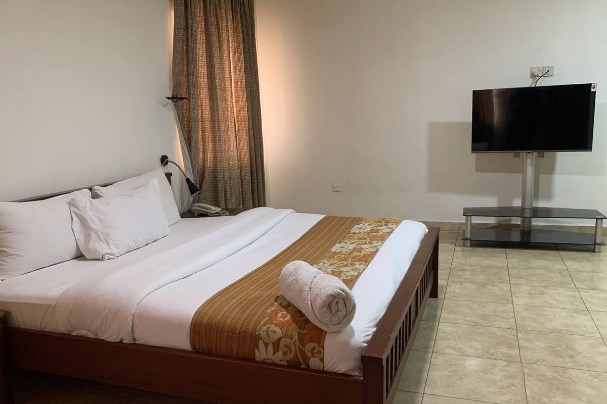 Una cama bien hecha con sábanas blancas y un plaid marrón decorativo, un televisor de pantalla plana sobre un soporte, y una cortina ligeramente abierta.