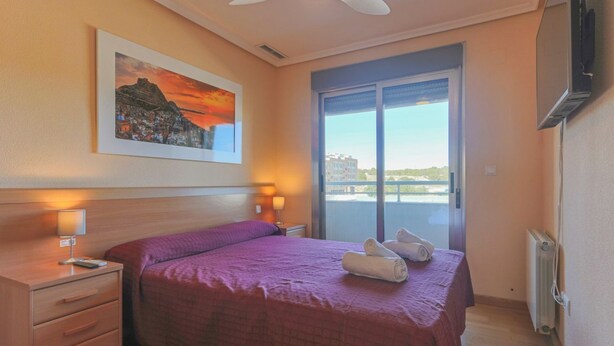 Een hotelkamer met een bed, nachtkastje, plafondventilator, raam met uitzicht en een schilderij aan de muur.