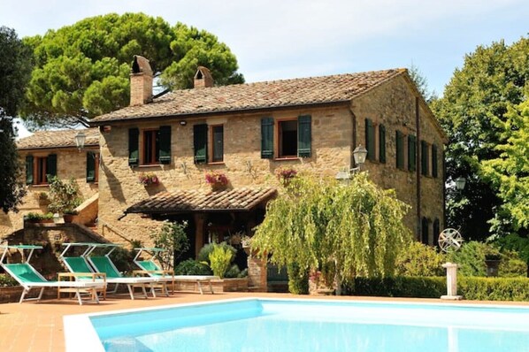 Un edificio de piedra con piscina, mobiliario exterior y un gran árbol.