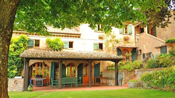Un espace extérieur abrité, doté de murs en pierre et d’un toit en tuiles, entouré d’une végétation luxuriante et avec un bâtiment en pierre en arrière-plan.