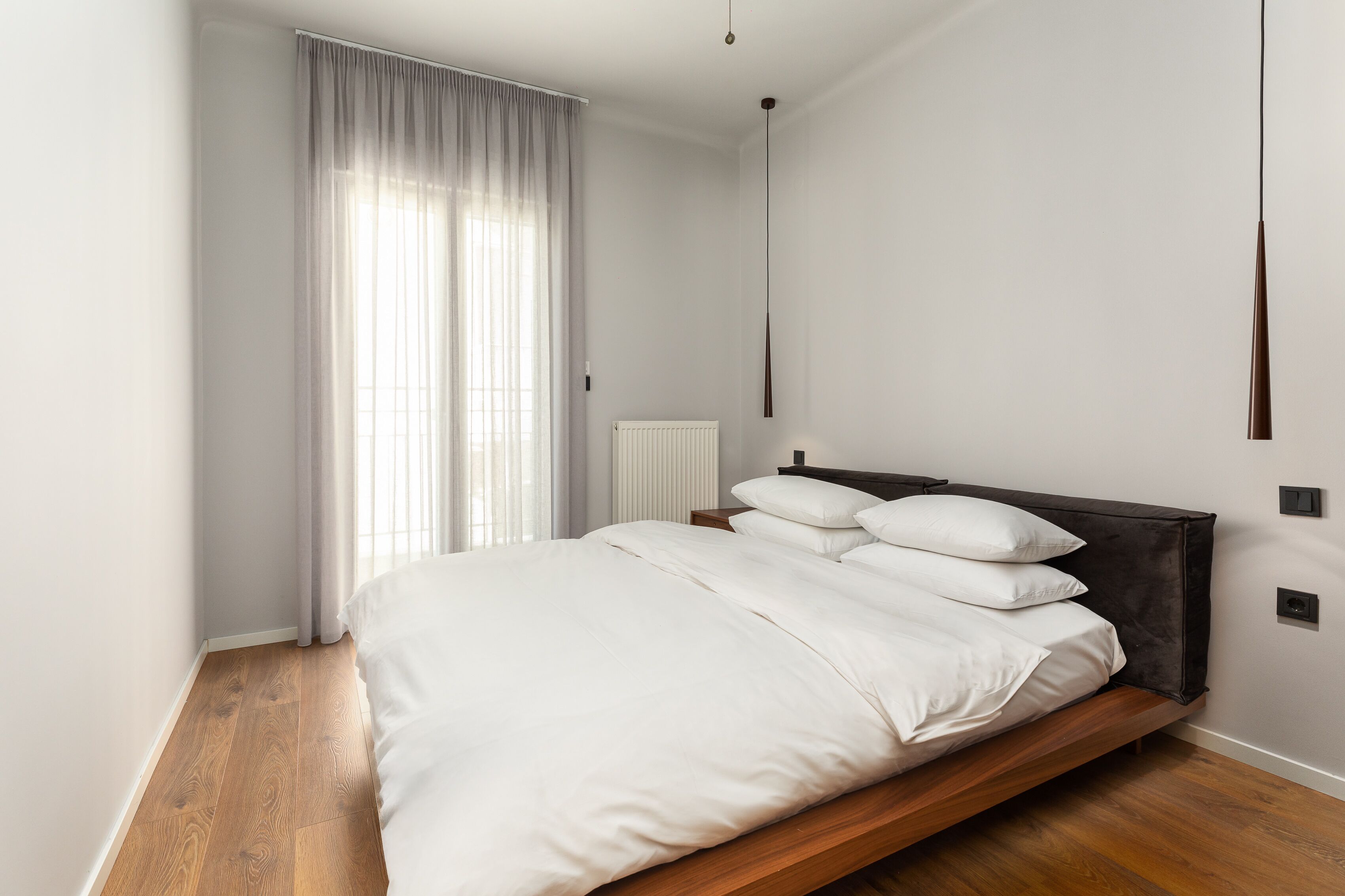 A modern bedroom with a wooden bed, white bedding, a radiator, and a window with sheer curtains.