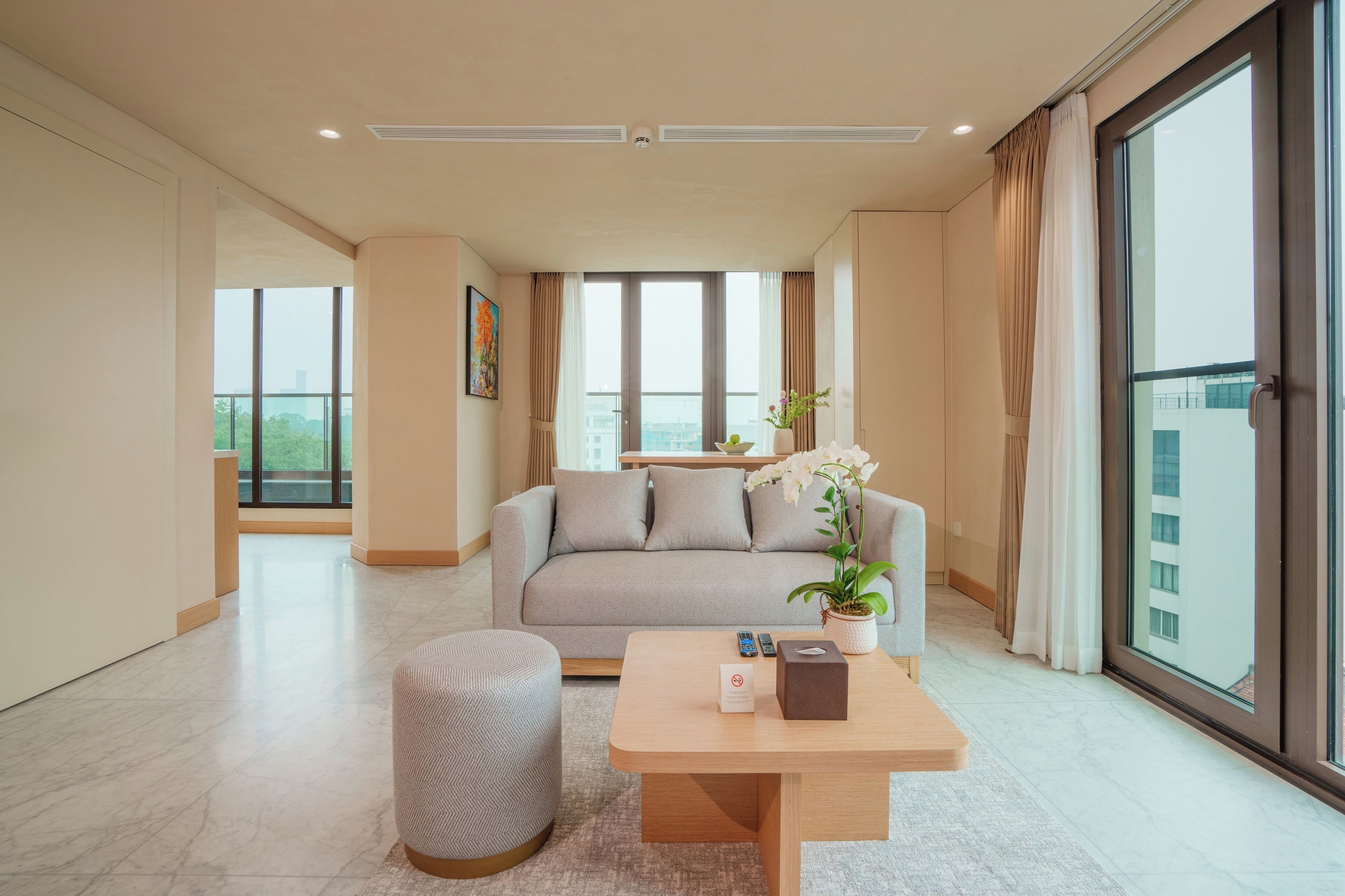 Un salon moderne avec un canapé, un pouf et une table basse, de grandes fenêtres et une vue sur les immeubles.