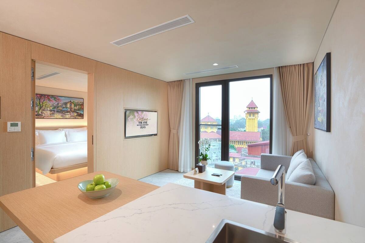 Una habitación de hotel moderna con cama, una zona de comedor con un bol de manzanas, un sofá y vistas a un edificio con una torre.