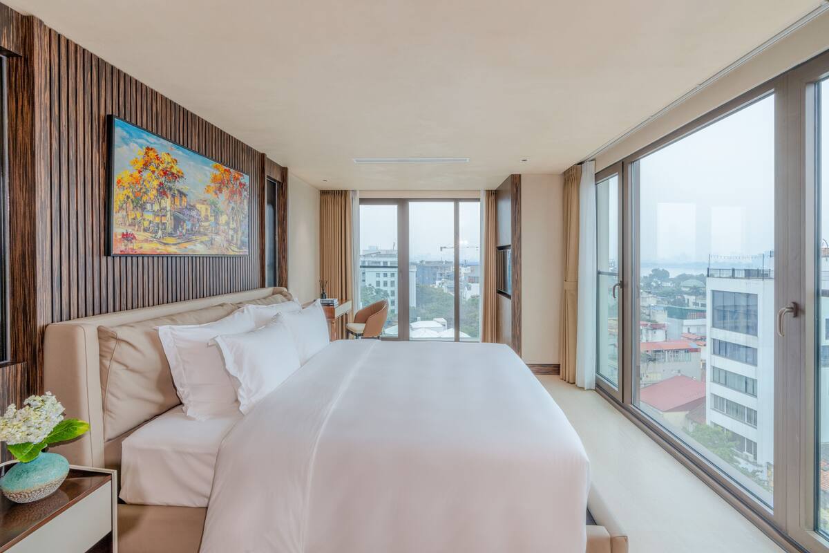 Habitación de hotel moderna con una cama grande, un cuadro en la pared y vistas de la ciudad a través de amplias ventanas.