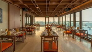Een ruime eetruimte met meerdere tafels en stoelen, grote ramen met uitzicht op de stad, en een houten plafond met rasterpatronen.