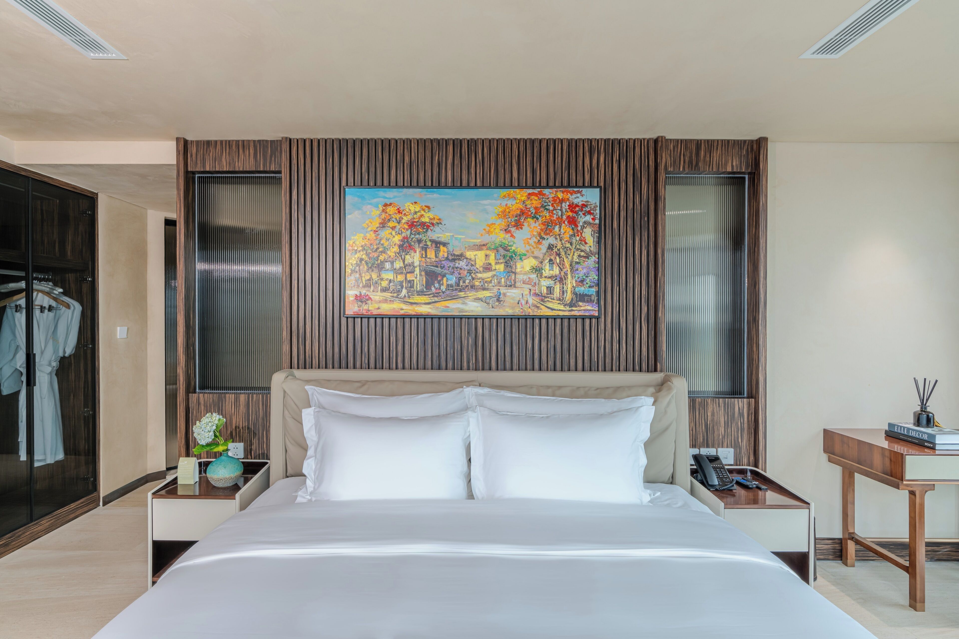 Une chambre d’hôtel moderne avec un grand lit, un tableau au-dessus du lit, une tête de lit en bois, une table de chevet avec une lampe et un bureau avec une chaise.