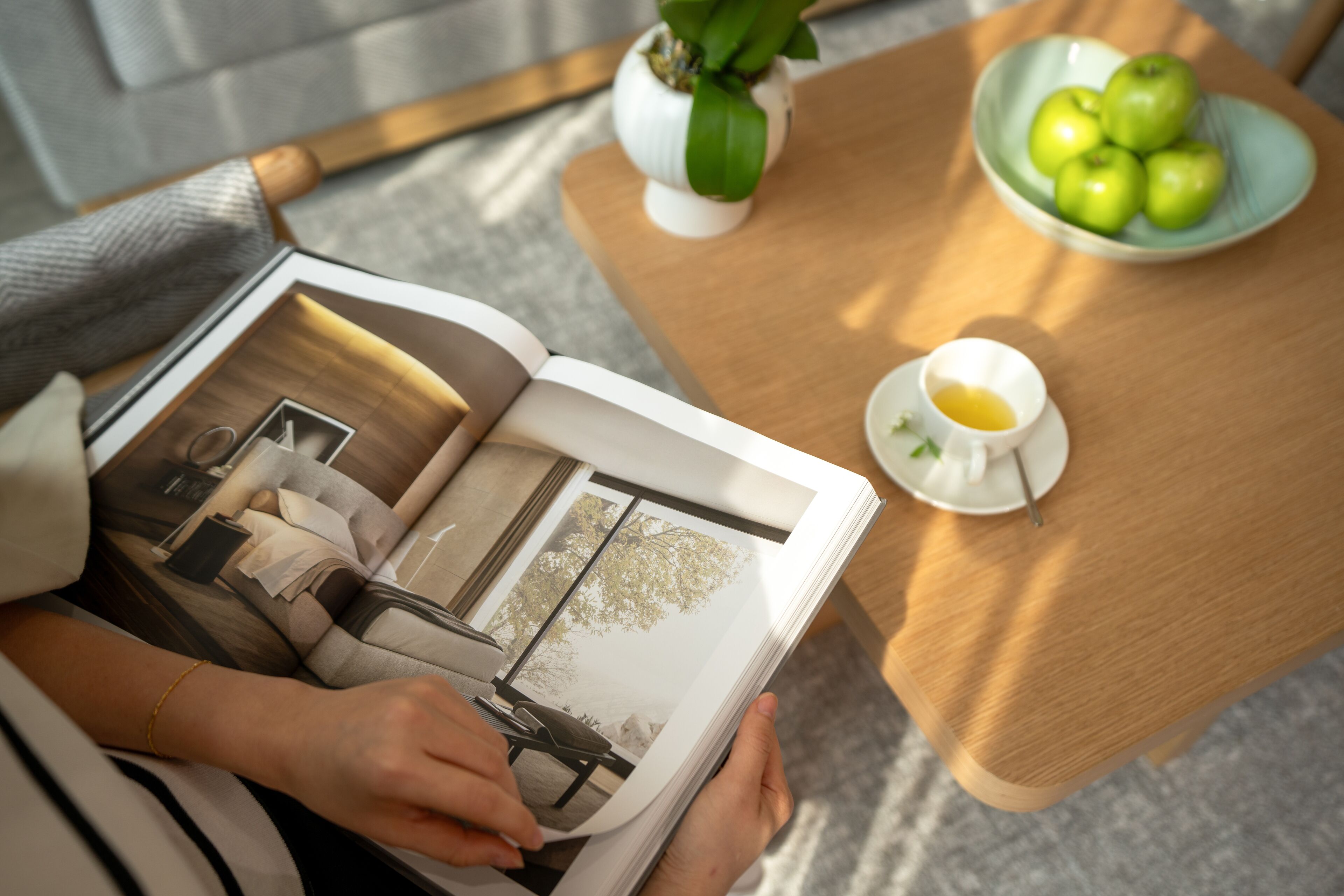 Una persona sostiene una revista con un diseño de cocina, una mesa de madera con un bol de manzanas verdes y una pequeña taza blanca con una cuchara.