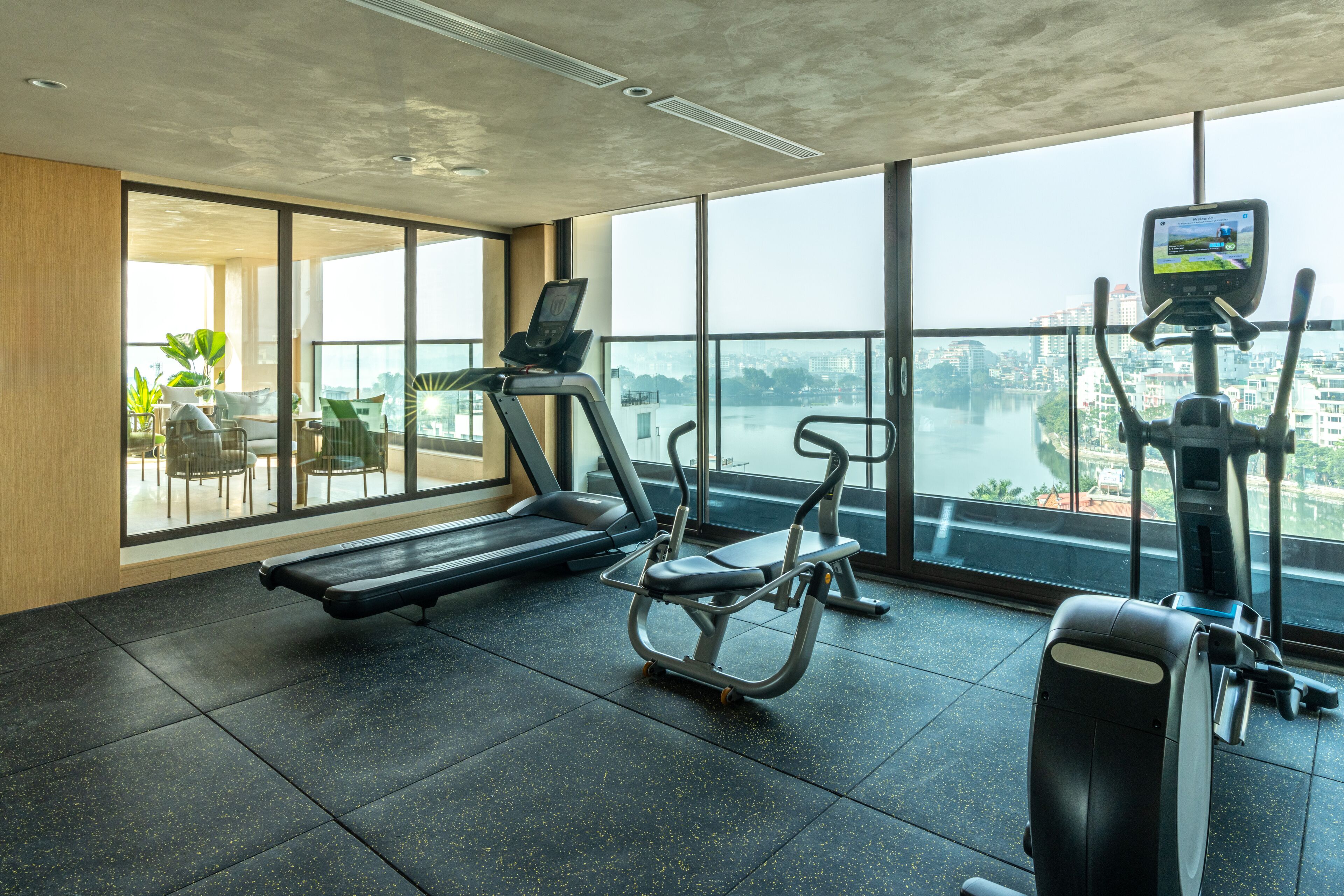 A gym with treadmills, an elliptical, and a rowing machine, with large windows offering a city view.