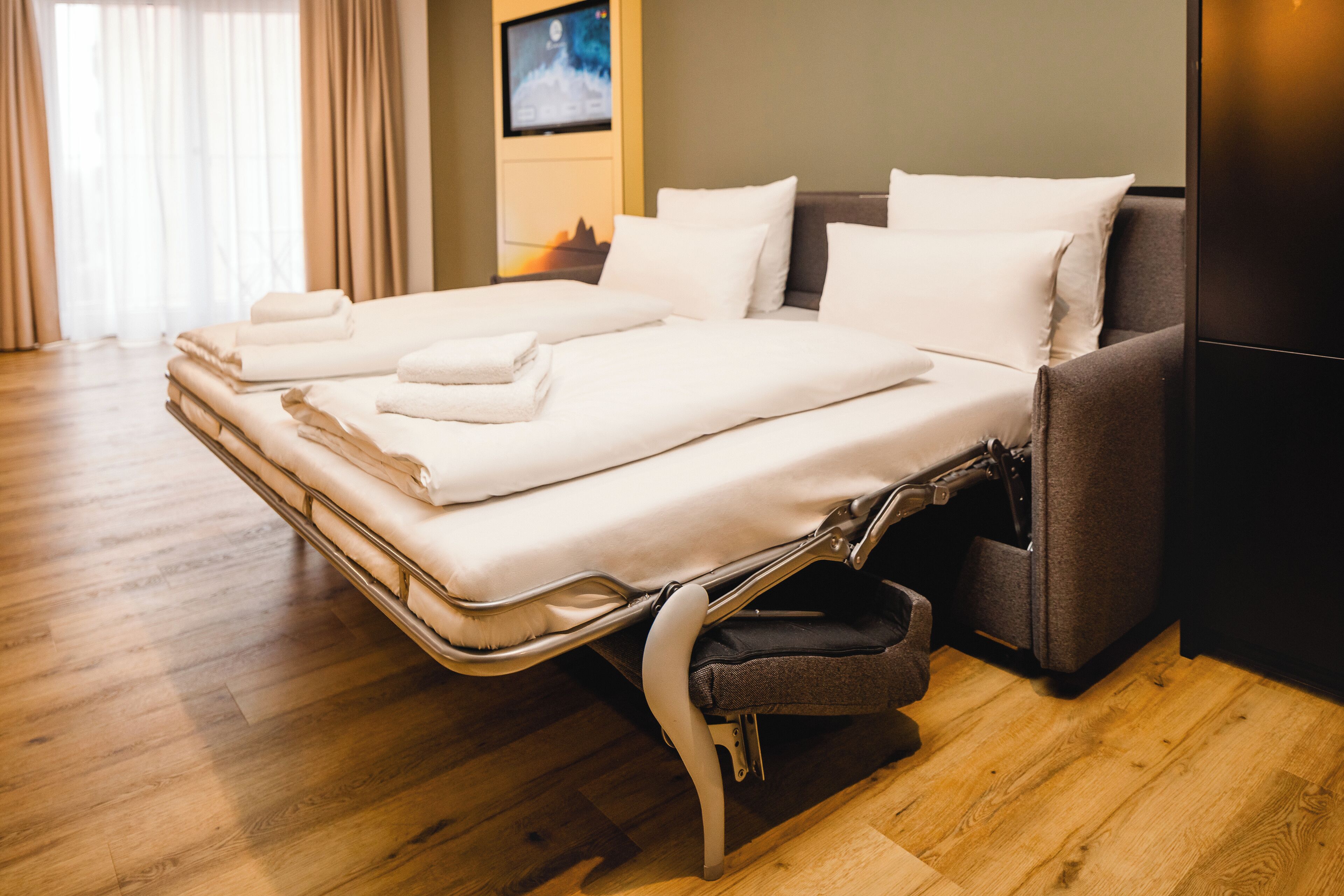 A neatly made bed with white linens and pillows, a grey sofa, and a wooden floor.