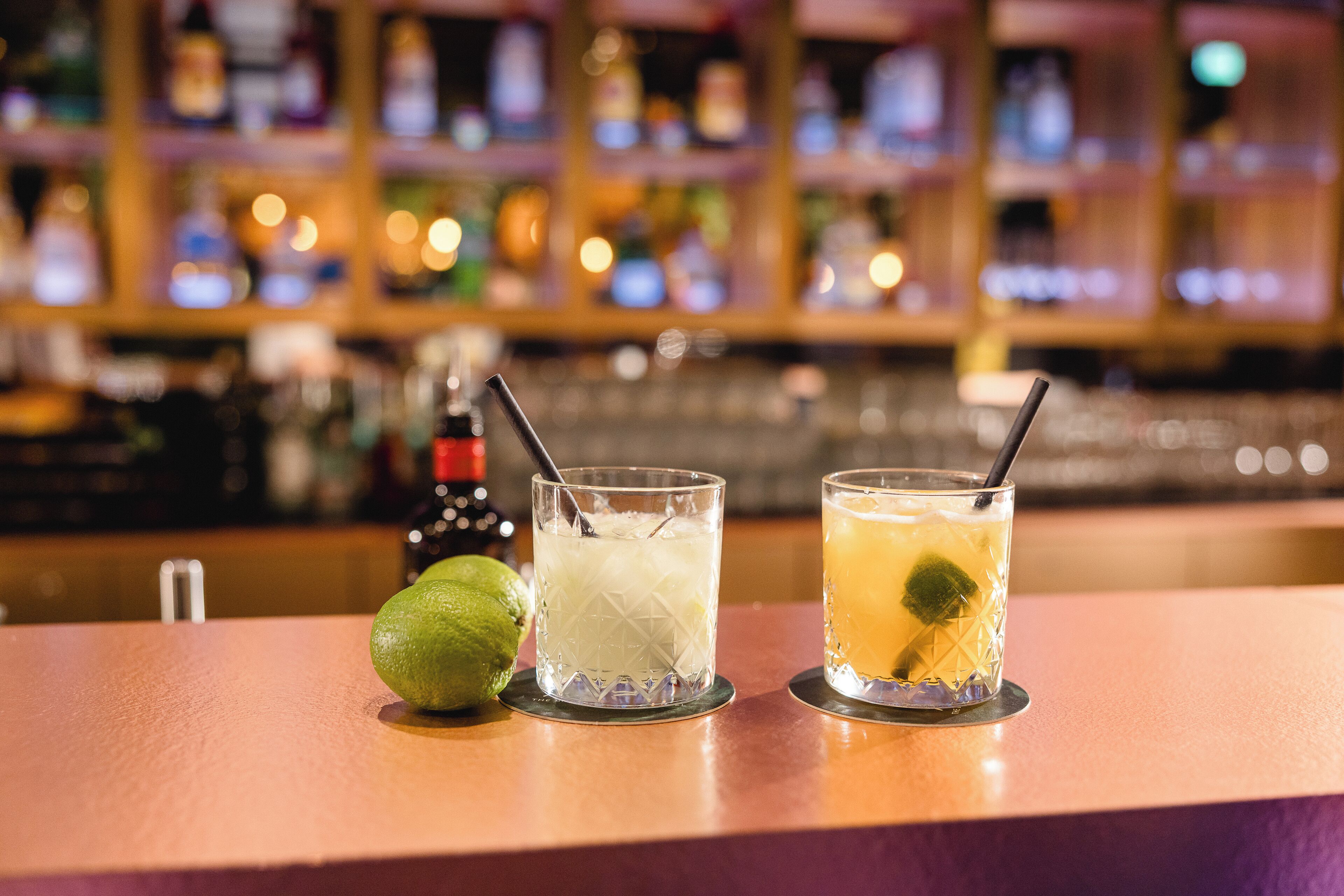 A bar counter with three glasses of beverages, a lime, and a bottle in the background.