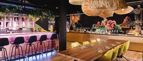 A modern bar area with a long wooden table, yellow chairs, and wicker hanging lights.
