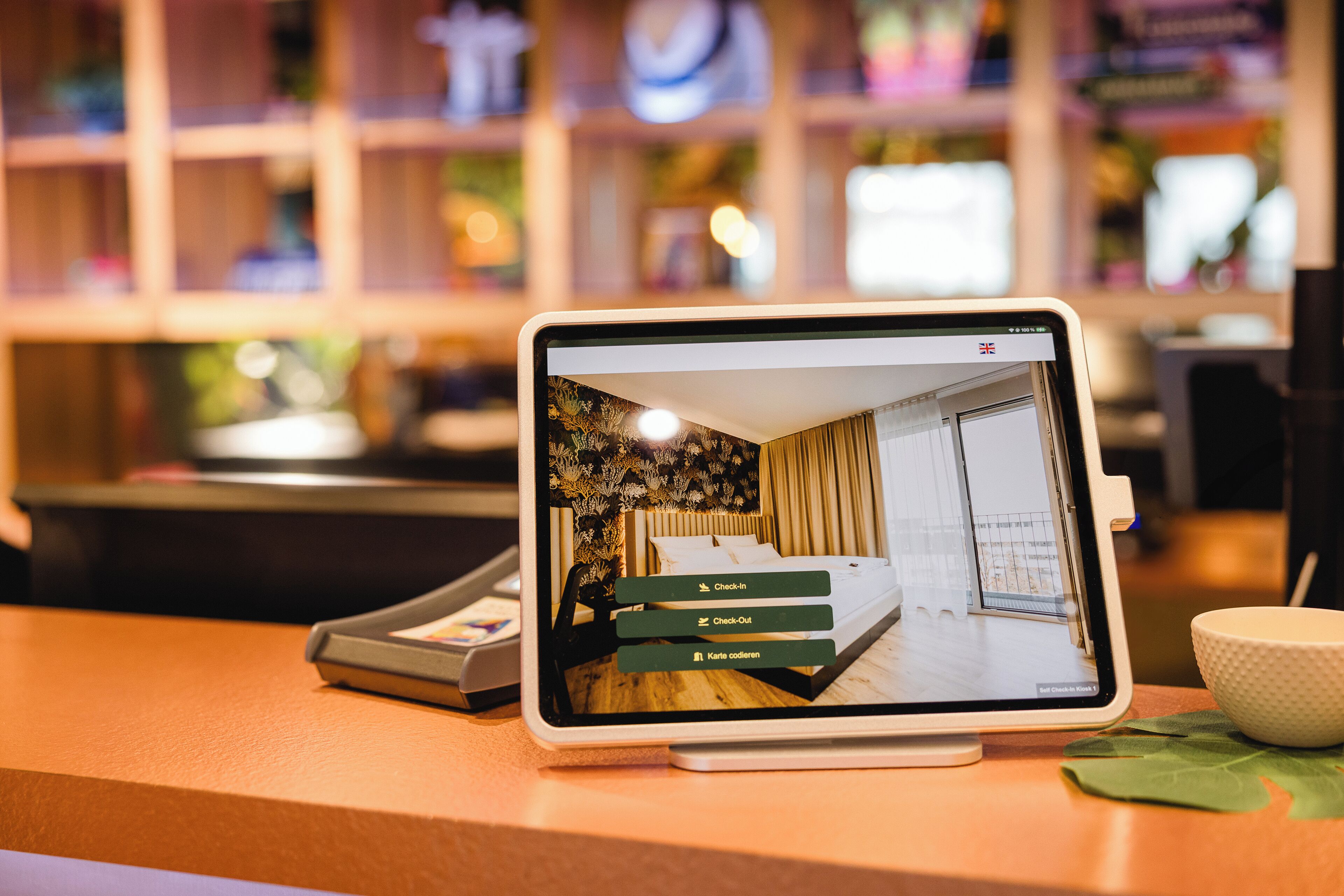 Een tablet toont een hotelkamer met een bed, een raam en een deur. Drie knoppen zijn zichtbaar, gelabeld 'Check-in', 'Check-Out' en 'Karte coden'.