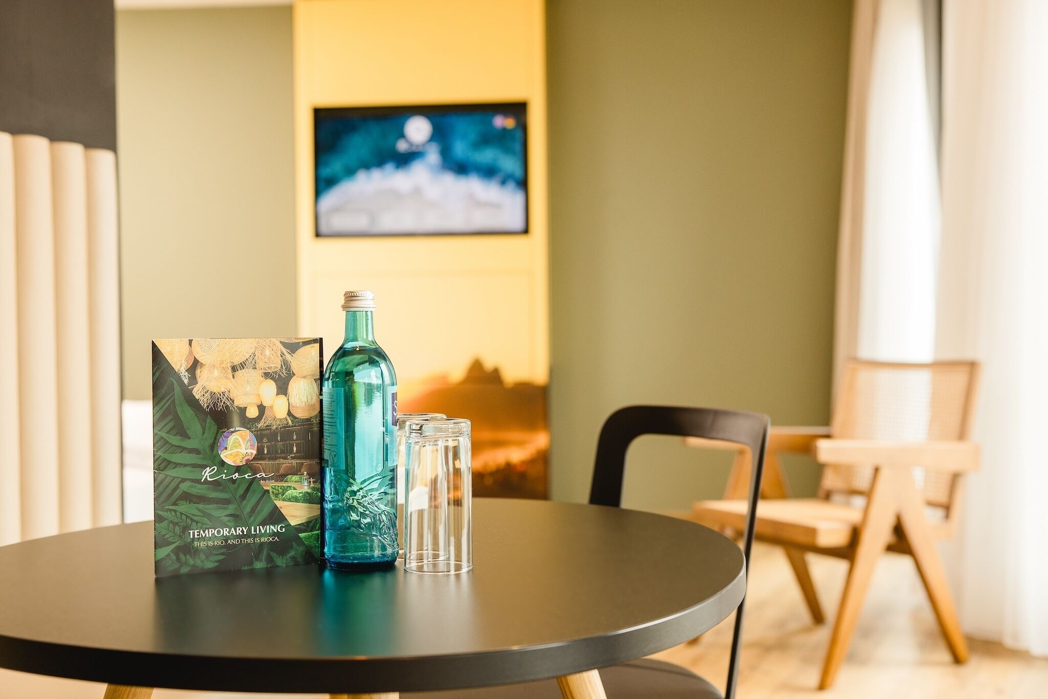 A table with a bottle and a glass, a framed picture, and a chair in a room with a wall-mounted TV.