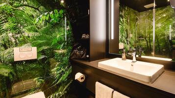 Un baño con un espejo grande, un lavabo y una ducha. Hay un perchero para toallas y un dispensador de papel higiénico. Las paredes están decoradas con plantas.