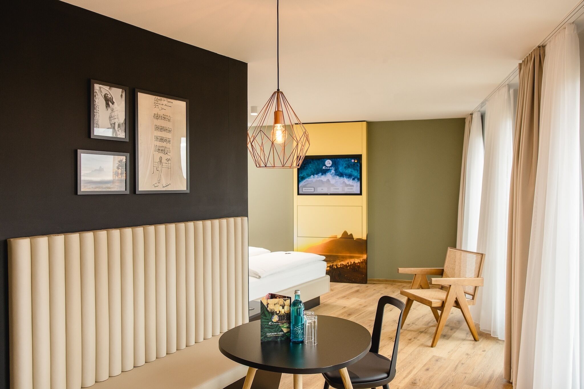Hotelkamer met een bed, een ronde tafel met een fles en glazen, een stoel, een tv aan de muur en ingelijste foto's.