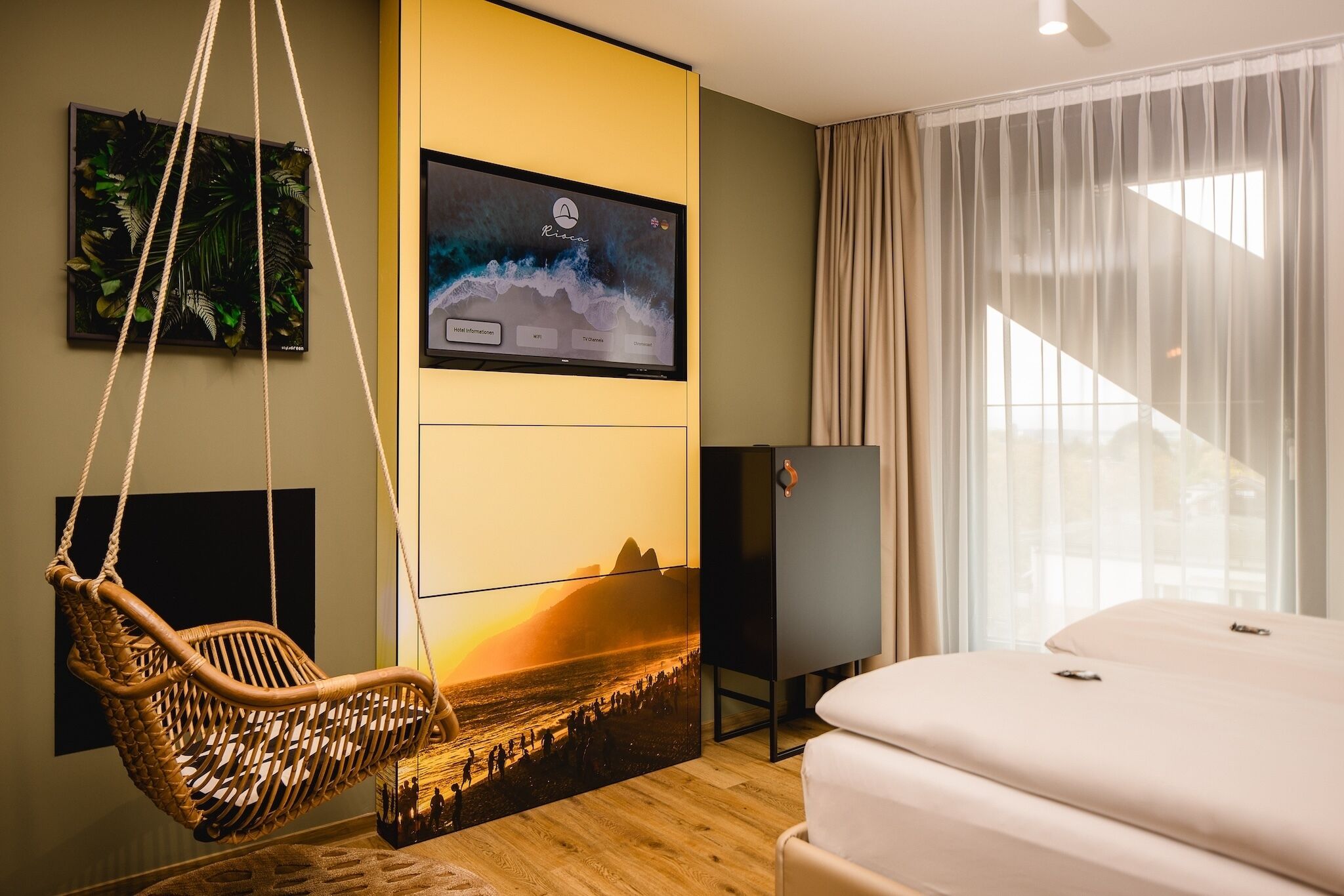 Et moderne hotelværelse med et stort fladskærms-tv på væggen, en hængestol, en seng og udsigt til det fri gennem lette gardiner.