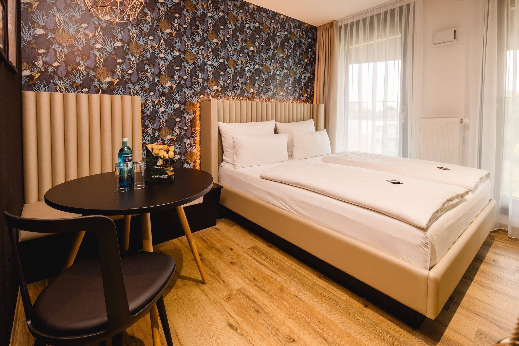 Habitación de hotel con cama, una pequeña mesa con sillas, un muro decorativo y una ventana con cortinas.
