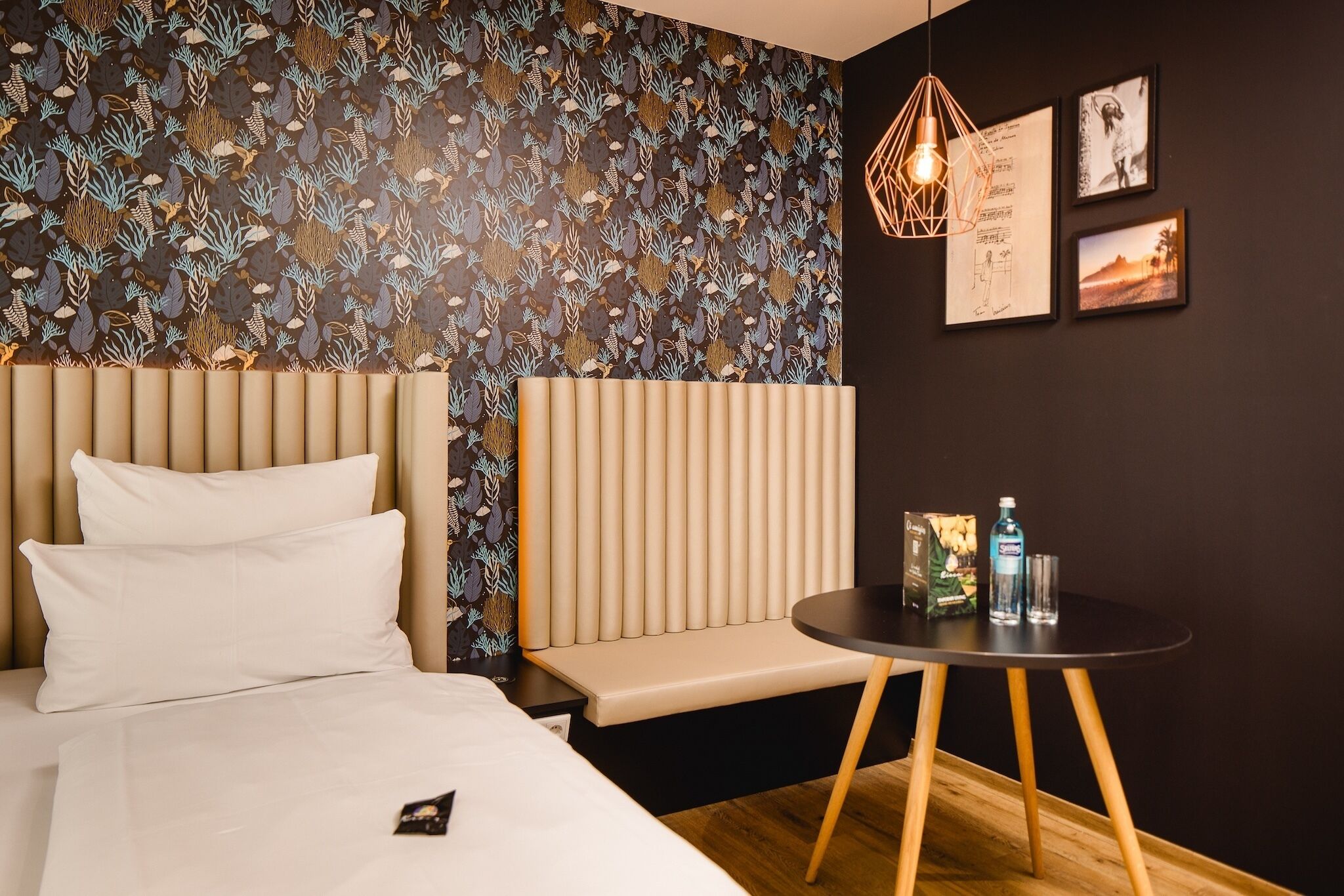 A hotel room with a bed, a bench, a small table, and a wall with a leaf-pattern wallpaper.