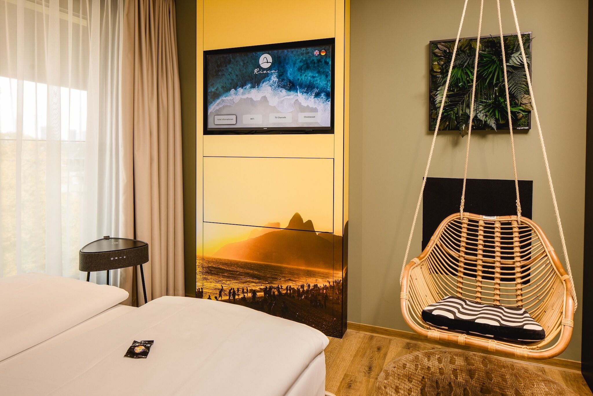 A hotel room with a bed, a TV mounted on the wall, a hanging chair, and a poster of a beach scene.