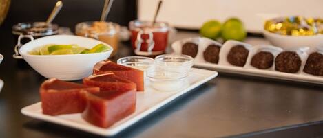 Una mesa con diversos alimentos, incluyendo un bol blanco con rodajas de lima, un plato con cubos rojos y una bandeja con bolitas de chocolate.