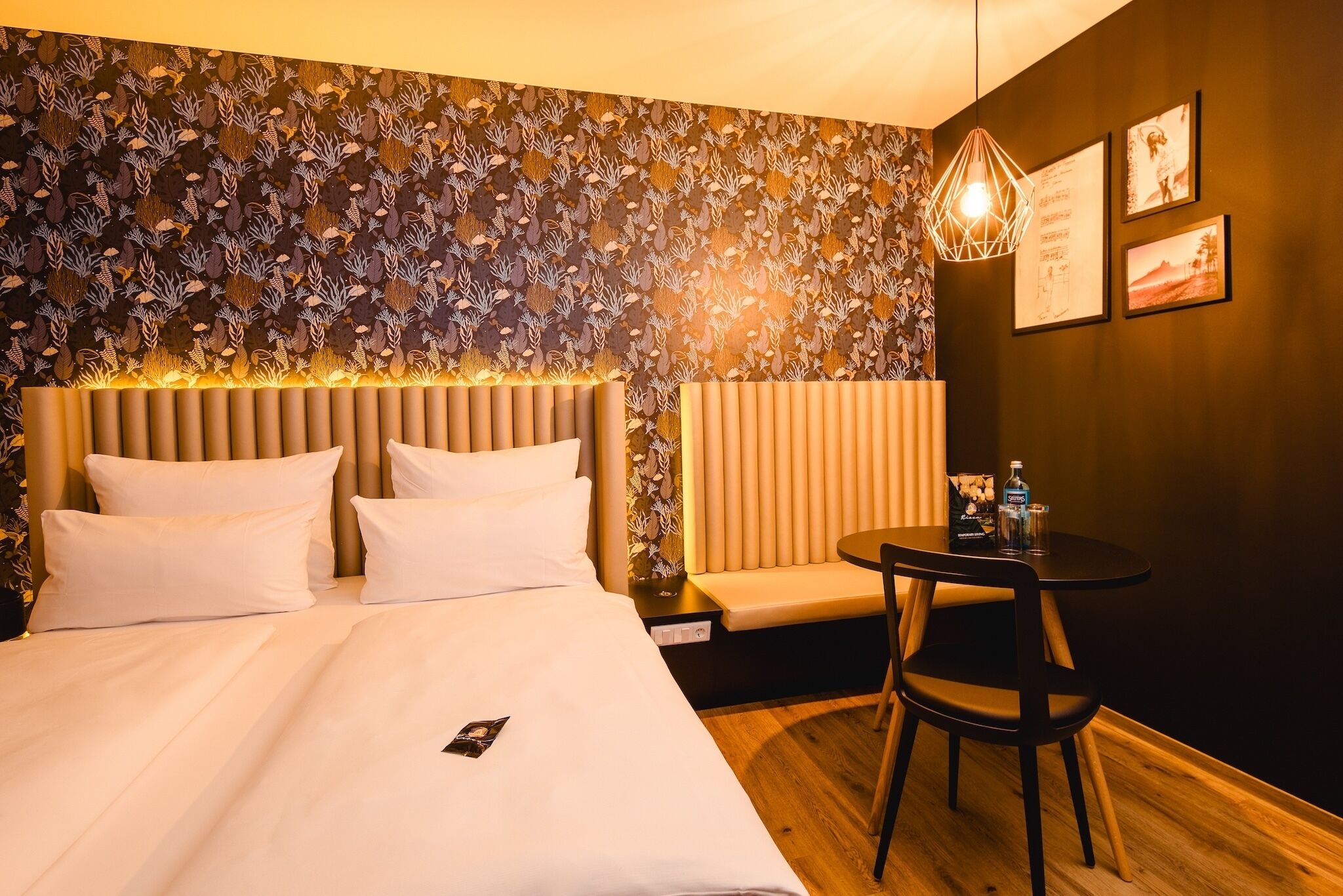 Hotellihuone, jossa on sänky, yöpöytä, tuoli ja seinä, jossa on kehystettyjä kuvia.