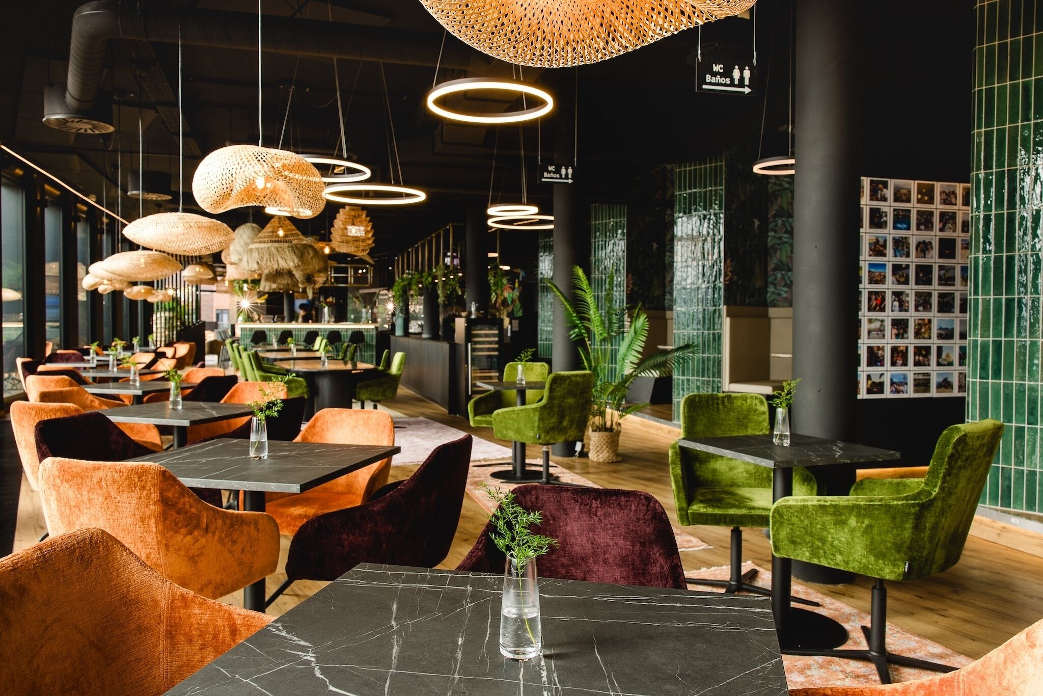 Een restaurantinterieur met meerdere tafels en stoelen, hangende lampen, en groene en oranje bekleden stoelen.