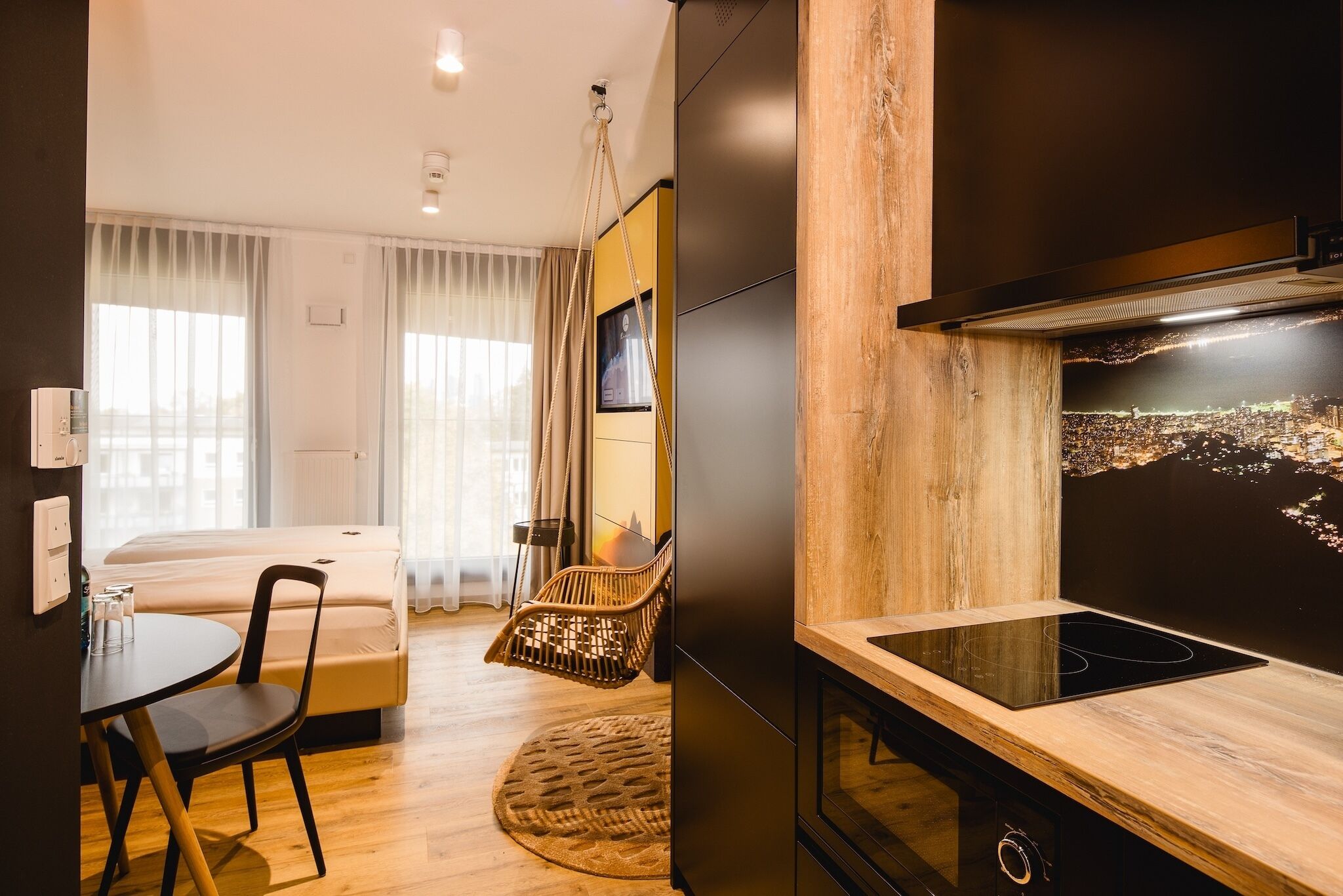 A modern hotel room with a bed, a dining area with a table and chairs, a kitchen with a stove, and a cityscape view on the wall.