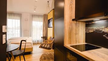 A modern hotel room with a bed, a dining area with a table and chairs, a kitchen with a stove, and a cityscape view on the wall.