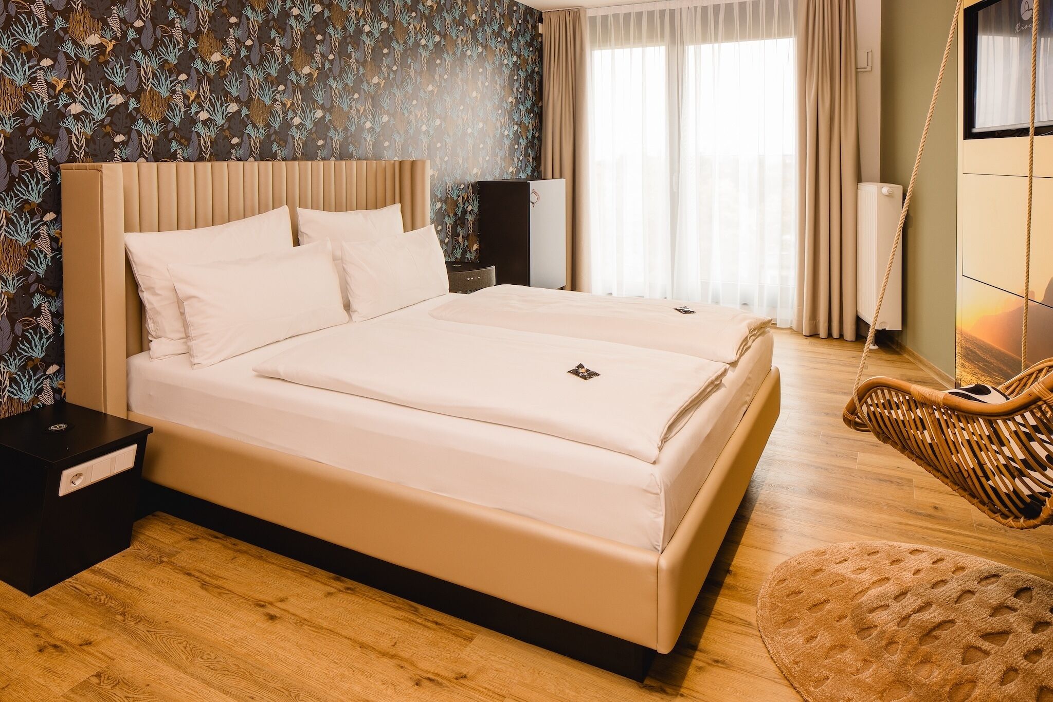 A double bed with a headboard, a bedside table, a hanging chair, and a patterned wall.