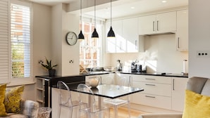 A modern kitchen with a black countertop, white cabinets, and a glass dining table with clear chairs. - The Chelsea Haven - Glamorous 1bdr Flat (London)