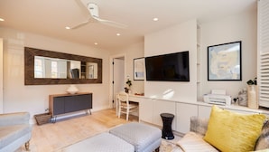 A modern living room with a sofa, ottoman, and a coffee table. There is a desk with a chair, a large mirror, and a flat-screen TV mounted on the wall. - The Chelsea Haven - Glamorous 1bdr Flat (London)