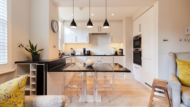 A modern kitchen with a central island, pendant lights, and a dining area with clear chairs. - The Chelsea Haven - Glamorous 1bdr Flat (London)
