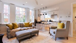 A modern living room with a sofa, ottoman, and armchair, a dining area with a table and chairs, and a kitchen with cabinets and a clock. - The Chelsea Haven - Glamorous 1bdr Flat (London)