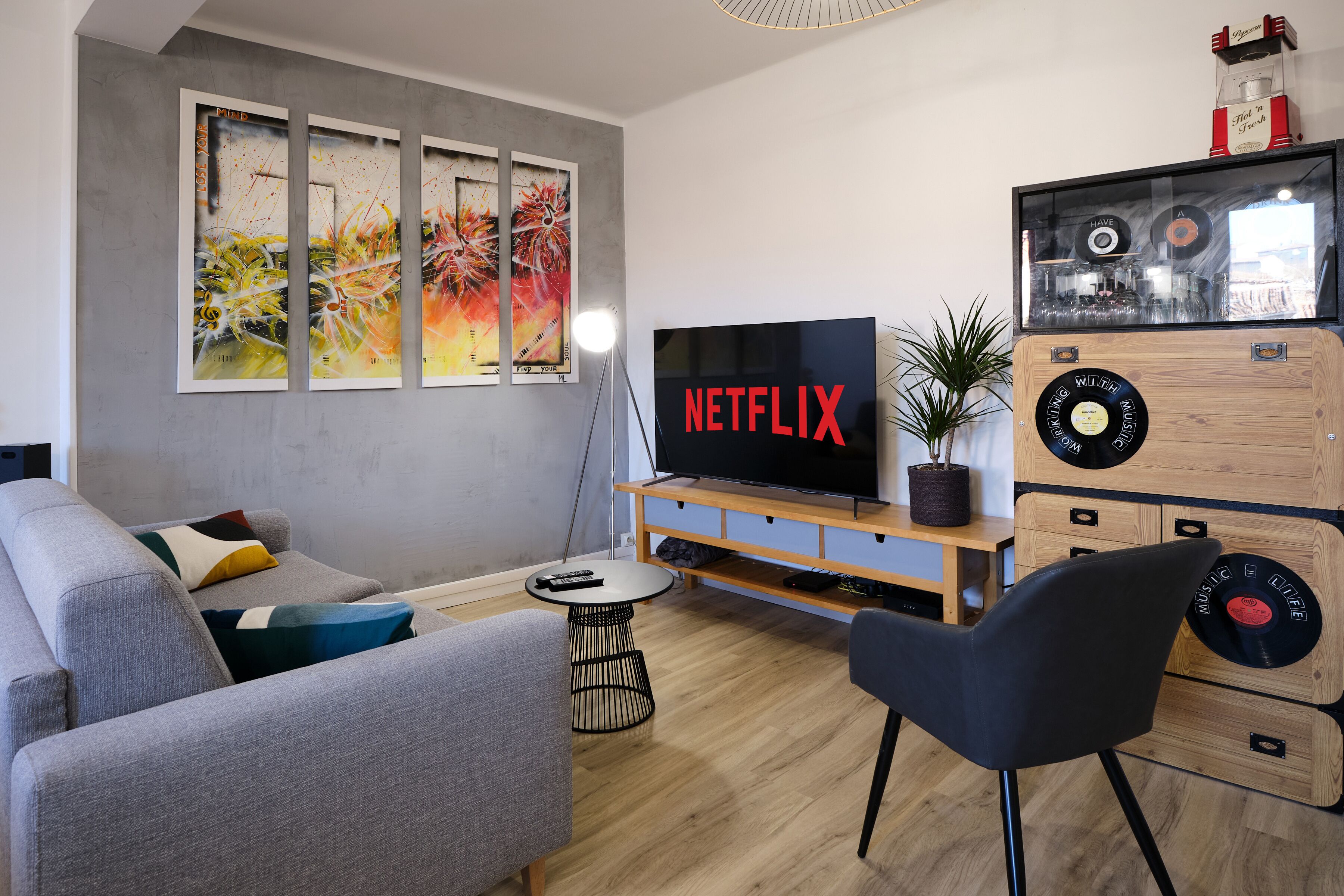 Una sala de estar moderna con un sofá gris, una silla negra, un mueble de madera para entretenimiento con un televisor mostrando 'NETFLIX' y una pared con arte abstracto.