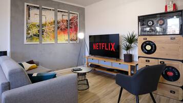 Una sala de estar moderna con un sofá gris, una silla negra, un mueble de madera para entretenimiento con un televisor mostrando 'NETFLIX' y una pared con arte abstracto.