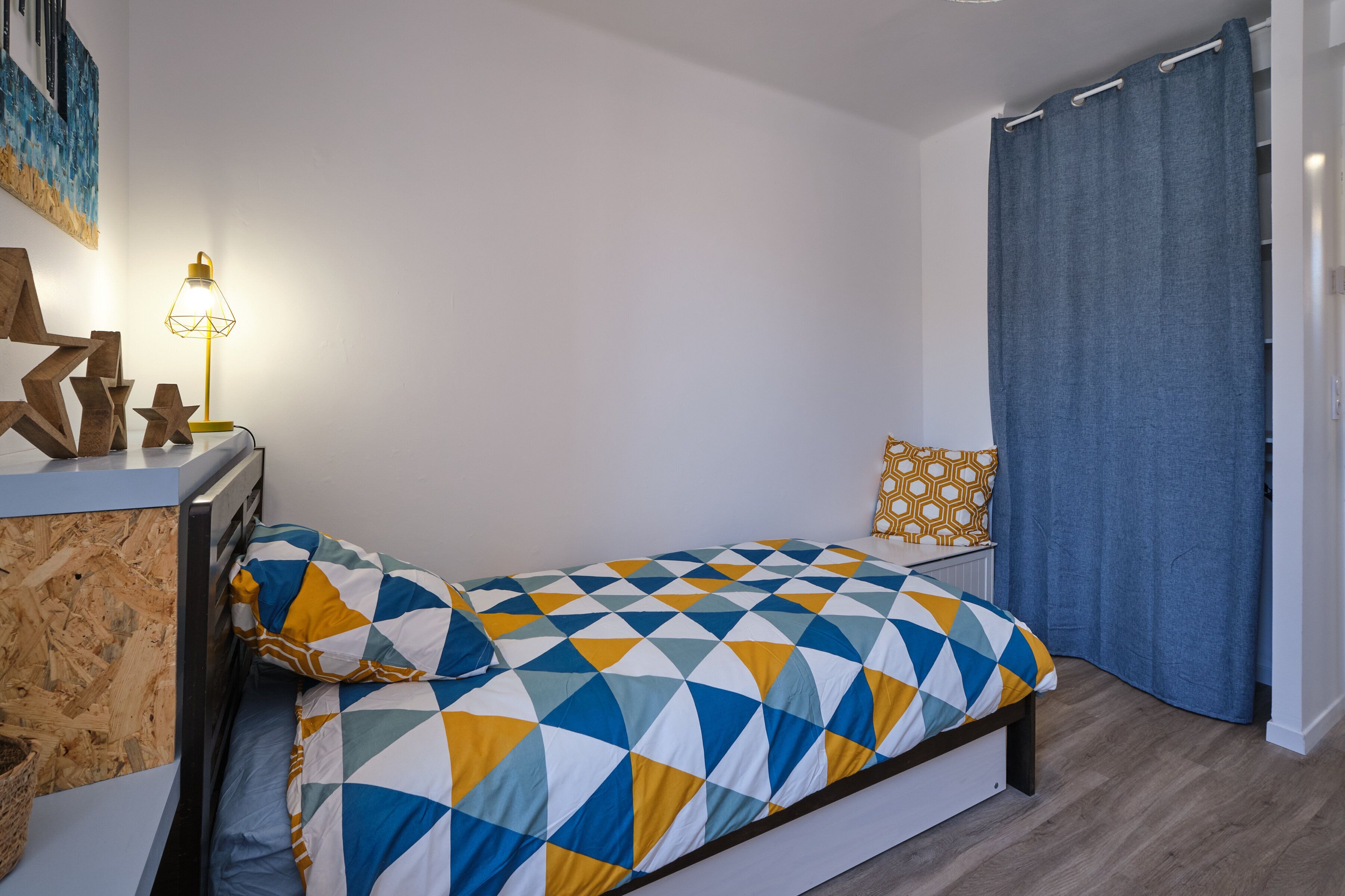 Un dormitorio con cama, mesita de noche, lámpara, ventana con cortinas y un diseño geométrico en la pared.