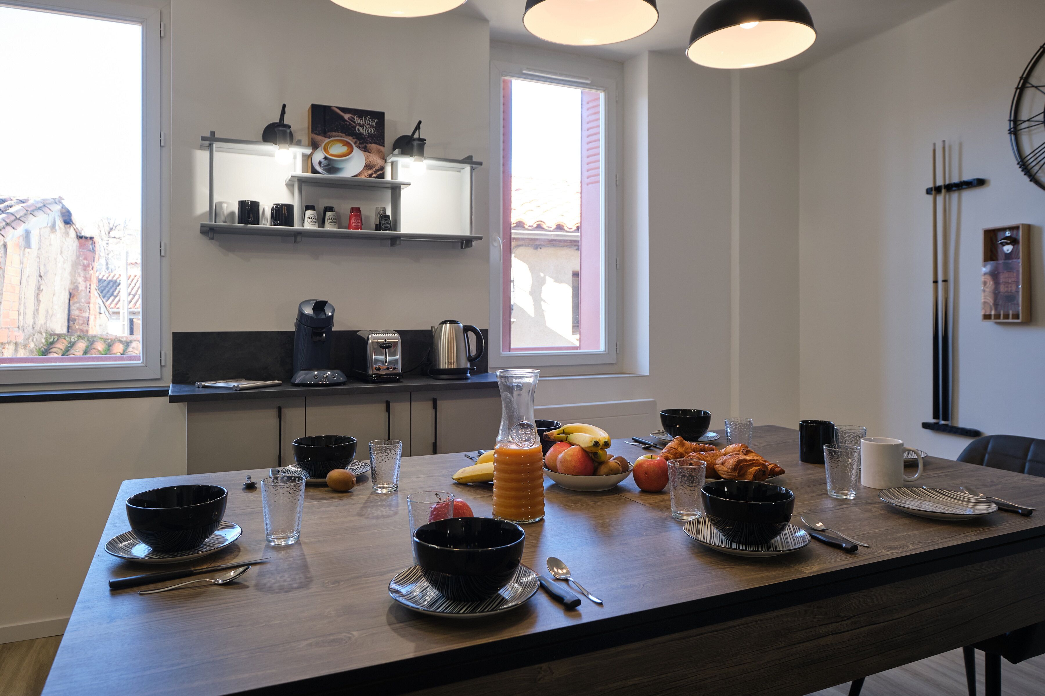 Une table à manger dressée avec des bols, des assiettes, des couverts et une carafe de jus d’orange.
