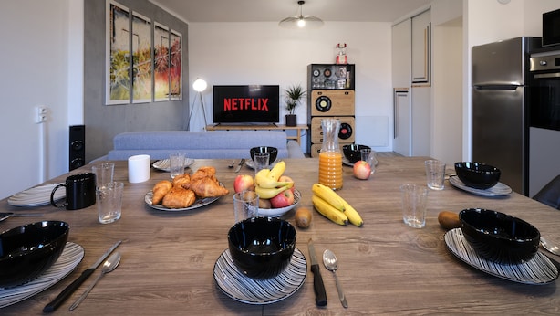 Una cocina moderna con una mesa de comedor dispuesta para el desayuno, un televisor mostrando 'NETFLIX', y una sala de estar con sofá y obras de arte.