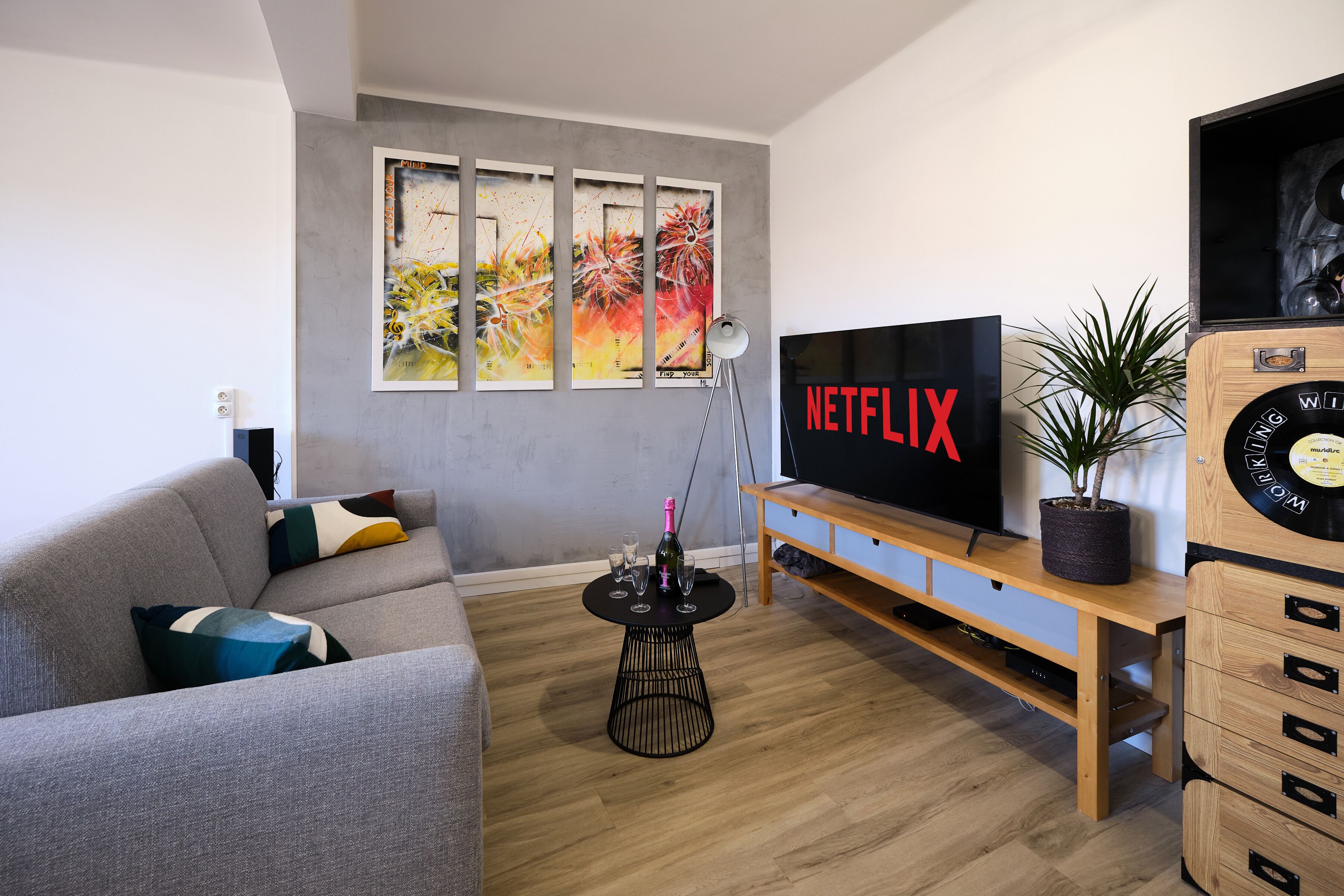 Una sala de estar moderna con un sofá gris, un mueble de televisión de madera, un televisor de pantalla plana grande y una obra de arte colgada en la pared.
