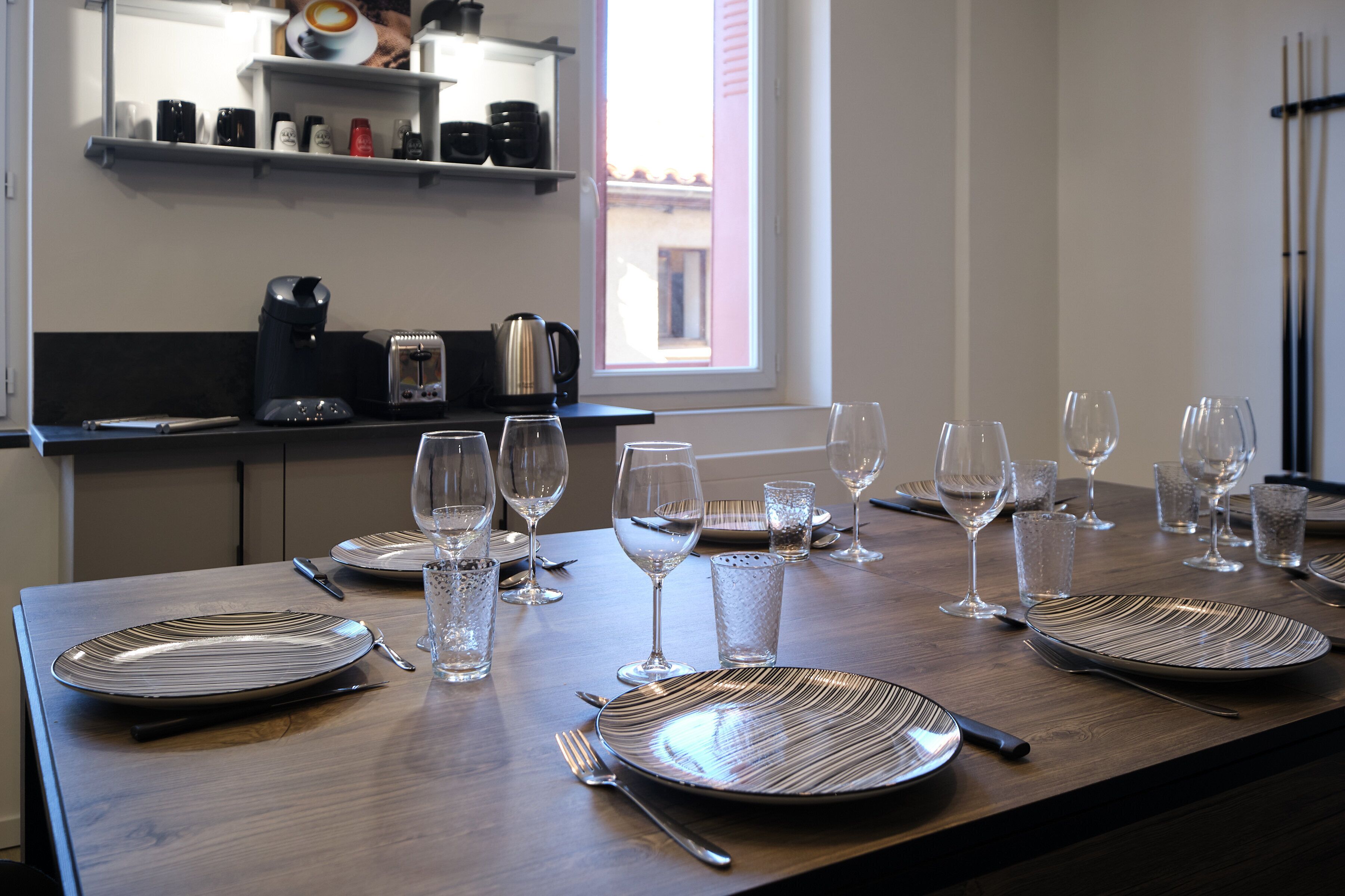 Una mesa de comedor dispuesta con platos, cubiertos y vasos, con una cafetera y un hervidor en la encimera.