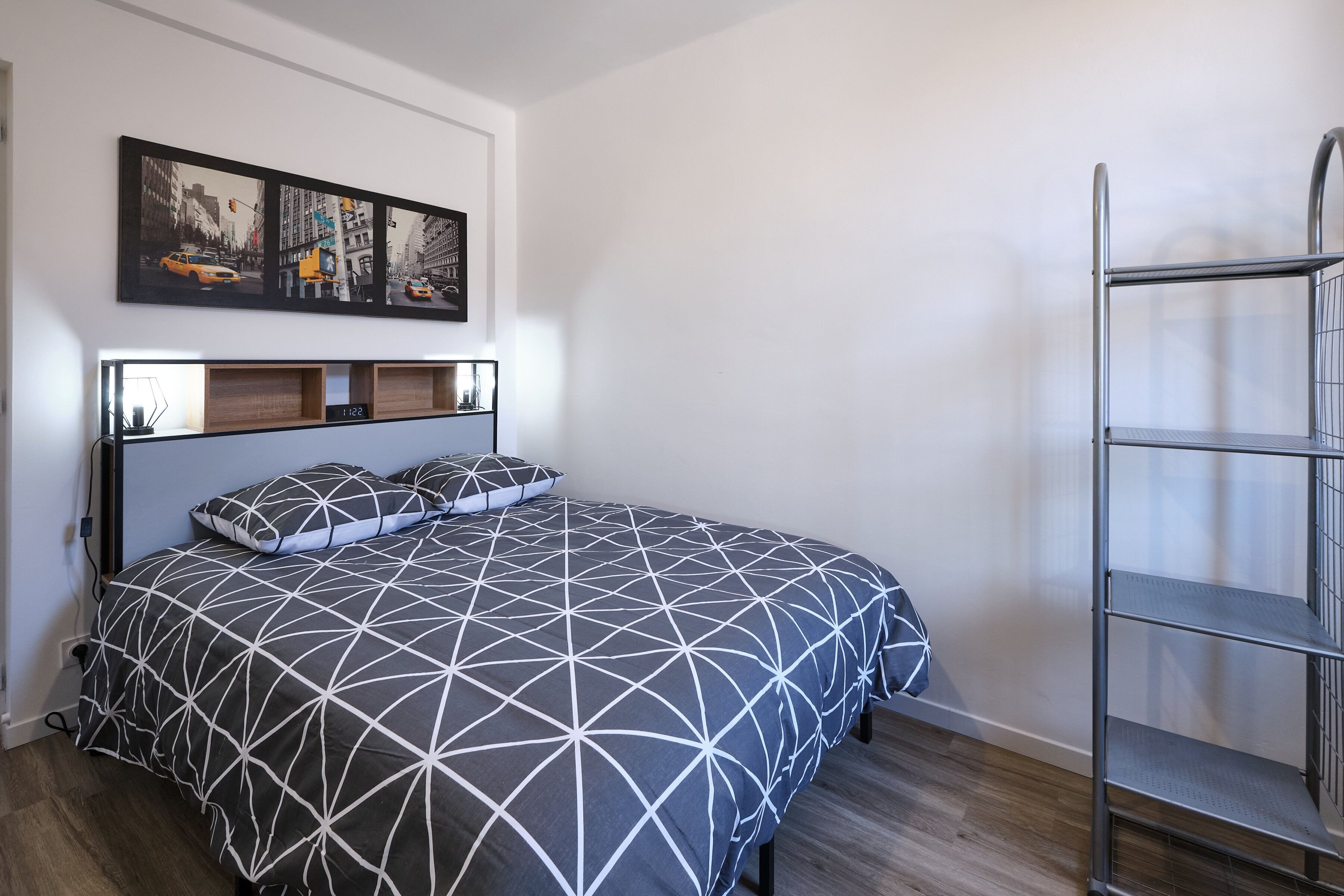 Ein ordentlich bezogenes Bett mit einem geometrisch gemusterten Überwurf, eine Wandablage mit einer Uhr und ein gerahmtes Stadtbild über dem Bett.