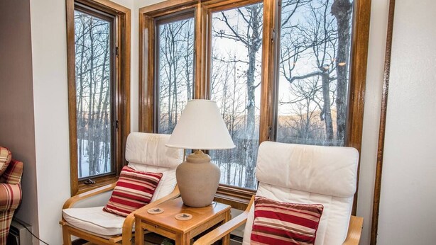 Rincón acogedor con una mesa de madera, dos sillas blancas con cojines, una lámpara y vistas a árboles sin hojas a través de amplios ventanales.