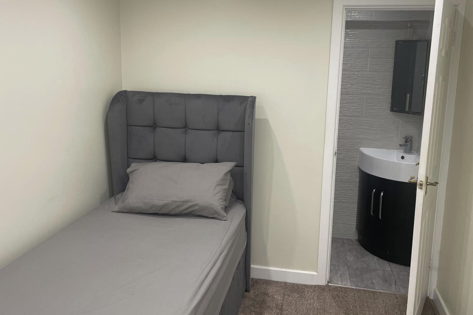 Una habitación pequeña con cama individual, cabecera tapizada de gris y colchón de color gris. Se ve un baño con lavamanos blanco y muebles de color oscuro a través de una puerta abierta.