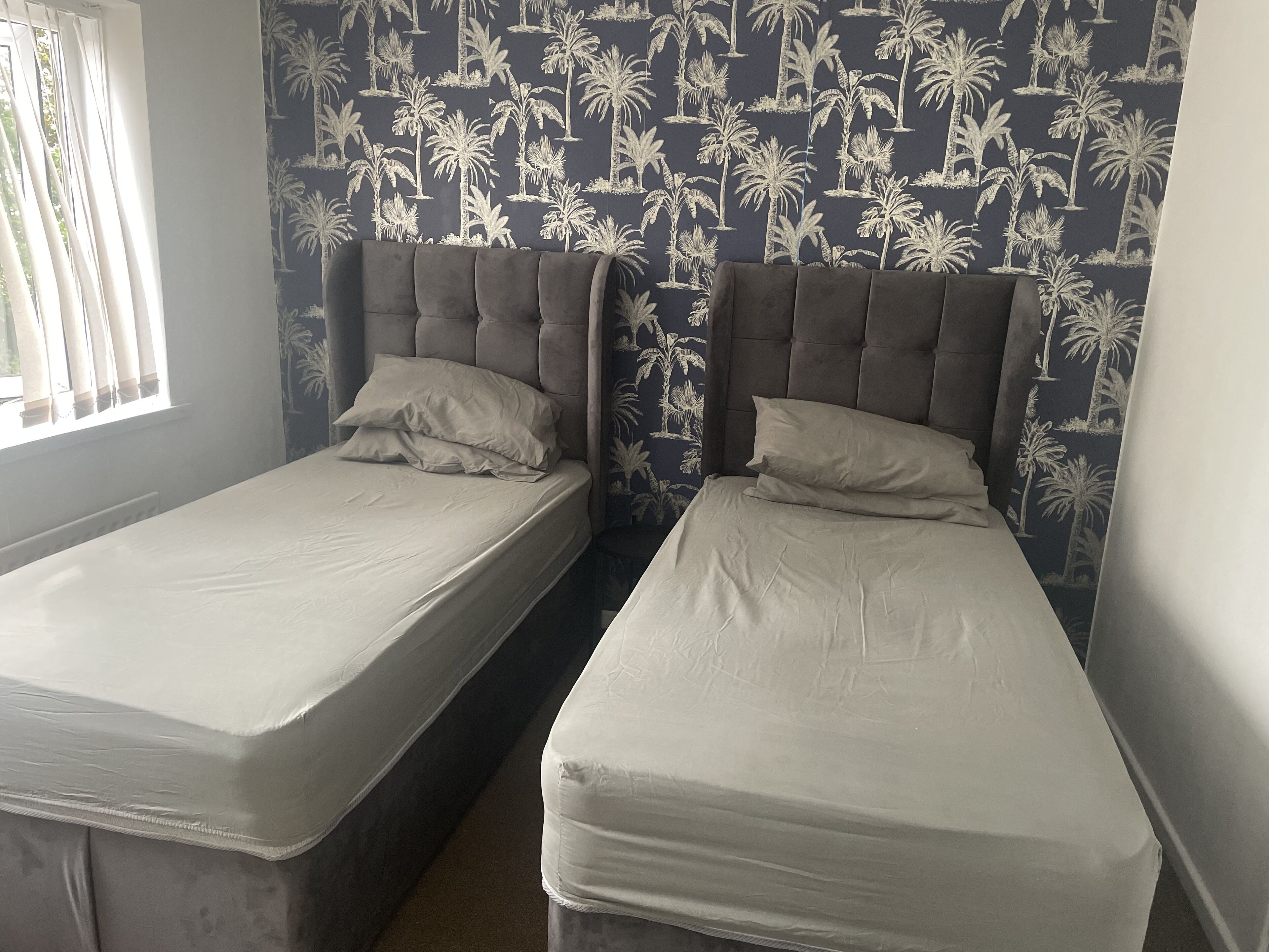 Two single beds with headboards, a window with curtains, and a wall with a palm tree pattern.